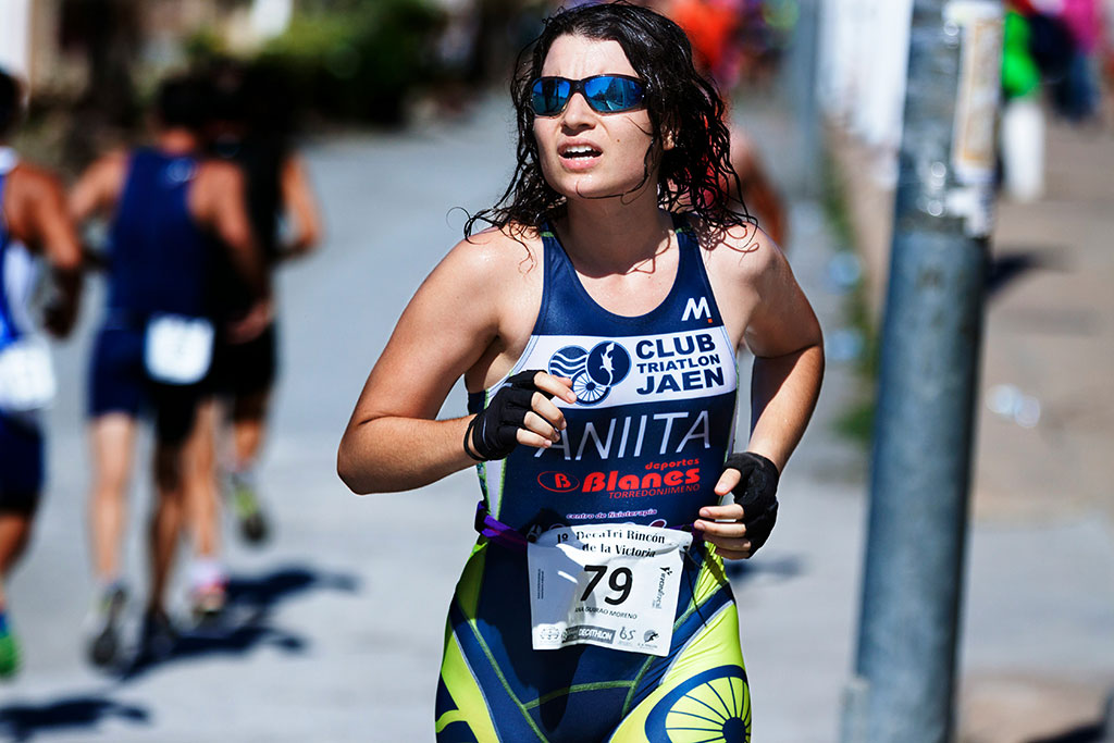 Image of a focused female triathlete running outdoors in bright sunlight while wearing sunglasses and athletic race gear.